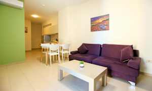 En lyst møbleret stue med en lilla sofa, et hvidt sofabord og en spiseplads ved siden af et køkken. (AI-genereret tekst)