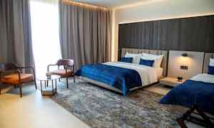 Et moderne hotelværelse med to senge, en siddegruppe med to stole og et bord samt mørke gardiner ved vinduet. (AI-genereret tekst)