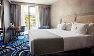 Et hotelværelse med en opredt dobbeltseng, et skrivebord med stol og vindue mod altan. (AI-genereret tekst)