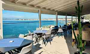 Udendørs servering med borde og stole nær havet, med udsigt over vandet og en by i baggrunden. (AI-genereret tekst)