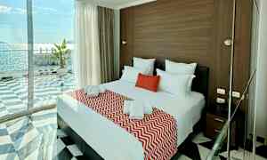 Et hotelværelse med en dobbeltseng dækket med røde og hvide tekstiler, store glasvinduer med havudsigt. (AI-genereret tekst)