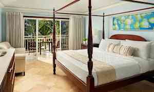 Rummet har en seng med fire stolper og er indrettet med lyse møbler. En balkon med udsigt over frodige palmer ses gennem de åbne glasdøre. (AI-genereret tekst)