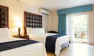 Hotelværelse med to senge, lyse vægge og mørke sengegavle. Bord med lampe og en altandør med udsigt til grønne omgivelser. (AI-genereret tekst)