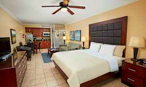 Et hotelværelse med en stor seng, natbord, polstret lænestol, spiseplads og et køkken i baggrunden. (AI-genereret tekst)