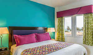 Et soveværelse med en dobbeltseng, farverigt sengetøj og puder. Væggene er turkise, og værelset har vinduer med udsigt over palmer og havet. (AI-genereret tekst)