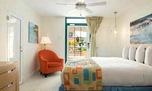 Et soveværelse med lys indretning. En seng med et farverigt sengetæppe, en orange lænestol, bordlampe og et vindue med udsigt ud. (AI-genereret tekst)