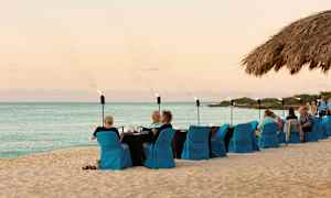 Strand med dækket bord og siddepladser med blå stolebetræk, hvor folk spiser og nyder selskab. Havet og himlen ses i baggrunden. (AI-genereret tekst)