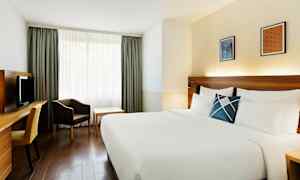 Et hotelværelse med en stor seng med hvide sengelinned, et skrivebord med stol, en lænestol og et bord ved et vindue med gardiner. (AI-genereret tekst)