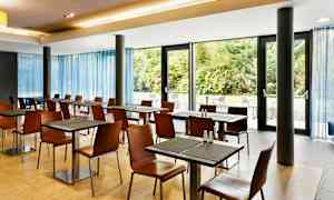 Et lyst spisestue med borde og stole ved store vinduer med udsigt til grønne buske. (AI-genereret tekst)