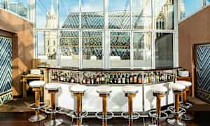 En cocktailbar med høje stole foran en glasvæg med udsigt over en bygning med farverige tagudsmykninger. (AI-genereret tekst)