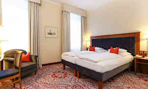 Et hotelværelse med to enkeltsenge, en mørk polstret sengegavl og røde pyntepuder. Værelset har hvide vægge, store vinduer med lyse gardiner og et rødt mønstret tæppe. (AI-genereret tekst)