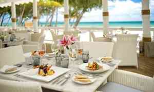 Et dækket restaurantbord nær stranden med tallerkener, glas og blomster. (AI-genereret tekst)