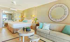 Et elegant hotelværelse med et stort spejl, en bred seng og en beige sofa med dekorative puder. Et rundt bord med mindre bord og plantepynt i midten. (AI-genereret tekst)