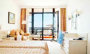 Et hotelværelse med to enkeltsenge, en sofa, et skrivebord og en balkon med udsigt. (AI-genereret tekst)