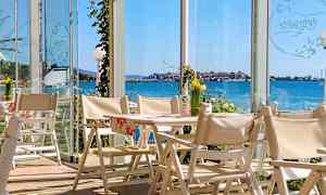 Udsigt fra en restaurant med hvide stole og borde, med glasvægge der giver udsigt over havet og en kystby. (AI-genereret tekst)