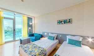 Et hotelværelse med to enkeltsenge, en blå lænestol og et bord. Værelset har lyse vægge og et stort vindue med gul gardin og balkon. (AI-genereret tekst)