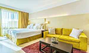 Et hotelværelse med en stor dobbeltseng, gul sofa, et bord med dekorativ vase og et rødt tæppe. (AI-genereret tekst)