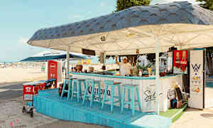 En strandbar med blå stole og tag, beliggende nær sandstranden. (AI-genereret tekst)