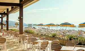 En udendørs café med borde og stole foran en strand med liggestole og parasoller. (AI-genereret tekst)