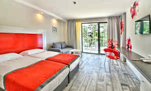 Et hotelværelse med to enkeltsenge, en sofa, en skrivebordsplads og en altan med udsigt til grønne omgivelser. (AI-genereret tekst)