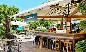 En café med borde og stole udendørs ved Sunshine Café, med grønne områder og havudsigt i baggrunden. (AI-genereret tekst)