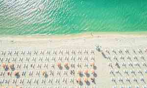 Luftfoto af en strand med parasoller og liggestole. Havet er flot grønblåt. (AI-genereret tekst)