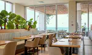 En restaurant med borde, stole og planter ved store vinduer med udsigt over havet. (AI-genereret tekst)
