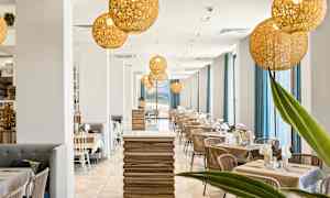 En moderne restaurant med en lys indretning med træborde, stole og store vinduer med blå gardiner. Dekorative belysningsarmaturer hænger fra loftet. (AI-genereret tekst)