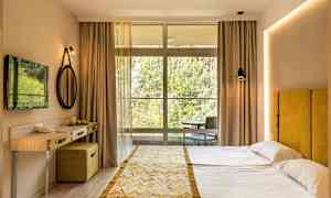 Et hotelværelse med en dobbeltseng, gul sengegavl, lille skrivebord og en stor vinduesfront mod naturen. (AI-genereret tekst)