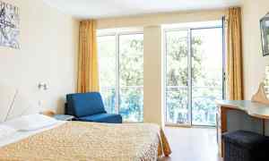 Værelse med seng, blå lænestol og skrivebord, med store vinduer ud mod altan. (AI-genereret tekst)