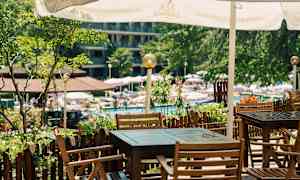 En udendørs terrasse med borde og stole af træ under en parasol, ved en have og poolområde. (AI-genereret tekst)