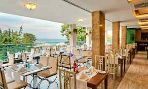 En restaurant med borde og stole i lyse farver, udsigt over havet og omgivet af træer. (AI-genereret tekst)