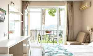 Et hotelværelse med en seng, et bord og en stol samt en balkon med havemøbler og udsigt til naturen. (AI-genereret tekst)