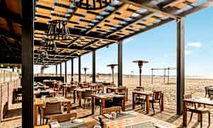 Udendørsrestaurant med træborde og stole, beliggende ved en sandstrand. (AI-genereret tekst)