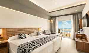 Et hotelværelse med to senge og en siddegruppe ved siden af en glasdør, der fører til en balkon med udsigt. (AI-genereret tekst)