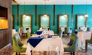 Et elegant restaurantinteriør med dækkede borde og grønne stole, omgivet af turkise vægge og dekorative hængelamper. (AI-genereret tekst)