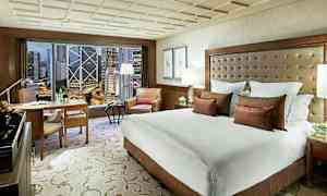 Et hotelværelse med en stor seng, lænestol og skrivebord. Vinduet viser en bymiljø med høje bygninger. (AI-genereret tekst)