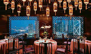 Et elegant indrettet restaurantlokale med runde borde, stole og lamper i loftet, med udsigt over en by gennem vinduerne. (AI-genereret tekst)