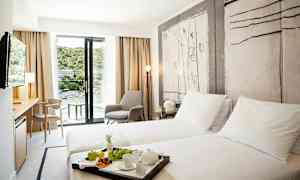 Et hotelværelse med to enkeltsenge, en siddegruppe og en altan med udsigt over et frodigt område. (AI-genereret tekst)