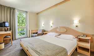 Hotelværelse med en stor seng, en lænestol, et skrivebord med et TV og et vindue med udsigt til grønne områder. (AI-genereret tekst)