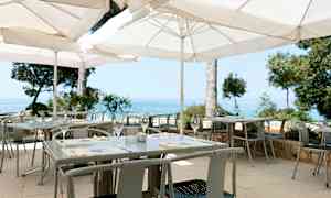 En udendørs servering med hvide parasoller, borde og stole samt udsigt over havet. (AI-genereret tekst)