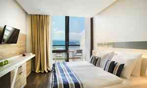 Et hotelværelse med en dobbeltseng, sengetøj i blå- og hvidstribet mønster og en stor glasvæg, der viser en balkon med udsigt over havet. Værelset har også et TV og skrivebord. (AI-genereret tekst)