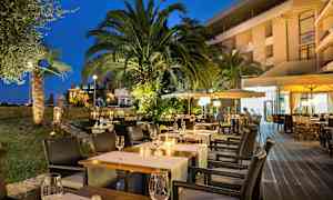 Aftenbillede af en udendørsrestaurant med oplyste borde, omgivet af palmer og grønt. (AI-genereret tekst)