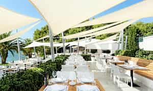 Et udendørsområde med flere borde og stole under spændte parasoller nær vandet. (AI-genereret tekst)