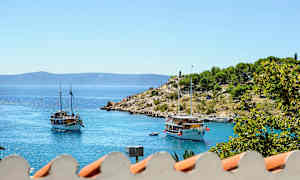 Billedet viser to sejlbåde på et roligt og blåt hav med en lille odde i baggrunden dækket af grønt. I forgrunden ses dele af et tag. (AI-genereret tekst)