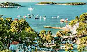 Udsigt over et poolområde med liggestole omgivet af frodig beplantning. I baggrunden ses en strand med både og en bugt med små øer. (AI-genereret tekst)