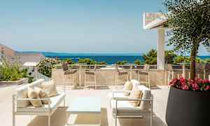 En terrasse med siddepladser og borde samt udsigt over havet. Den er omgivet af planter og træer med klar himmel. (AI-genereret tekst)