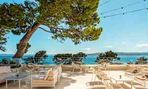 Udendørs terrasse med stole og borde, med udsigt til havet og de omkringliggende træer. (AI-genereret tekst)