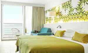 Et hotelværelse med grønt sengetøj, vægdekorationer og en siddegruppe ved et vindue mod altanen. (AI-genereret tekst)