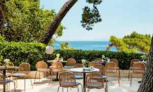 Billede af en udendørsservering med borde og stole, omgivet af grønt og med havudsigt. (AI-genereret tekst)